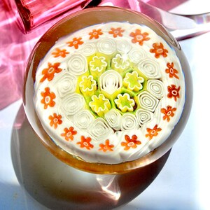 May include: A glass paperweight with a white background and a circular pattern of orange, yellow, and green flowers.