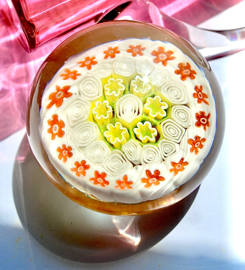 May include: A glass paperweight with a white background and a circular pattern of orange, yellow, and green flowers.