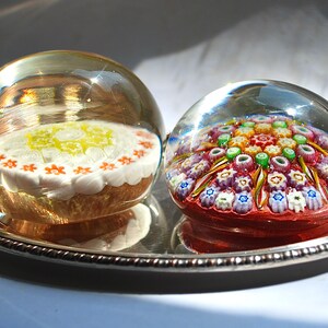 May include: Two glass paperweights on a silver tray. One paperweight features a white and yellow floral design, while the other features a colorful, multi-colored floral design.