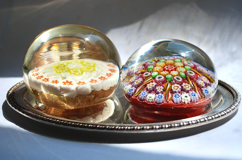 May include: Two glass paperweights on a silver tray. One paperweight features a white and yellow floral design, while the other features a colorful, multi-colored floral design.