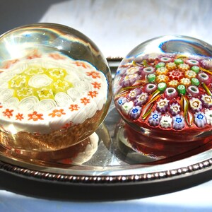 May include: Two glass paperweights with intricate floral designs. One paperweight features a white and yellow floral pattern, while the other has a multi-colored floral pattern.
