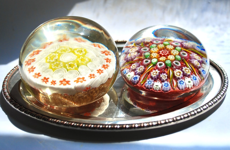 May include: Two glass paperweights with intricate floral designs. One paperweight features a white and yellow floral pattern, while the other has a multi-colored floral pattern.