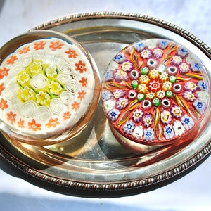 May include: Two colorful glass paperweights with intricate floral designs. One paperweight features a white background with orange and yellow flowers, while the other has a red background with multi-colored flowers.