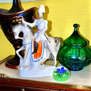 Op de afbeelding: Een verzameling decoratieve items op een tafel. Een wit keramieken beeldje van een man op een paard, een groene glazen pot met deksel, een glazen presse-papier met een blauw bloemmotief en een zwart-gouden lampvoet.