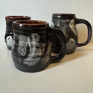 May include: Three handmade ceramic mugs with a dark charcoal gray glaze and a glossy finish. Each mug features hand-painted white ghost figures with simple black dot eyes scattered across the exterior surface and a brown interior rim.