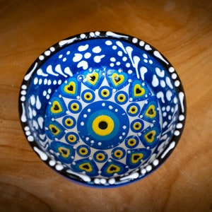 Puede incluir: Un pequeño cuenco de cerámica azul con un diseño blanco y amarillo. El cuenco presenta un motivo central de ojo con una pupila negra y un iris azul. El cuenco está rodeado por un anillo de puntos blancos y un anillo de corazones amarillos.