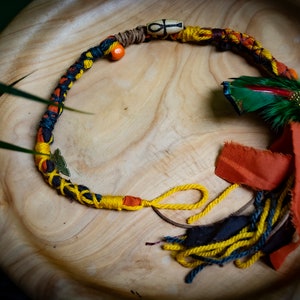 以下が含まれることがあります： 木製のビーズ、アンクのチャーム、羽根のアクセントが付いたカラフルな編み紐。紐は木製の表面に巻き付けられており、黄色、青、オレンジの糸のタッセルが付いています。