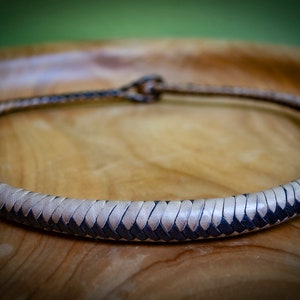 May include: A braided leather choker necklace. The necklace is a light tan color with a dark brown border. The necklace is circular and appears to be handmade. The background is a wooden surface.