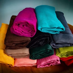 Op de afbeelding: Een verzameling opgevouwen stoffen sjaals in verschillende kleuren, waaronder geel, bruin, roze, blauwgroen, zwart, groen en grijs. De sjaals zijn gerangschikt in een houten kom, wat een aantrekkelijke textielpresentatie creëert.