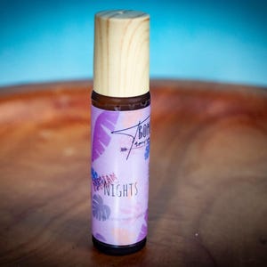 May include: A small, cylindrical essential oil bottle with a light wood cap. The label is lavender with tropical leaf and floral designs, and the words "Hawaiian Nights" are printed on the label.
