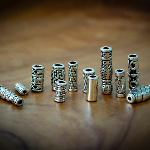 Puede incluir: Una colección de cuentas cilíndricas plateadas con varios patrones en relieve. Las cuentas tienen diferentes longitudes y diseños, incluyendo patrones florales, geométricos y texturizados. Están dispuestas sobre una superficie de madera marrón.
