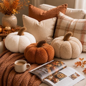 May include: Three plush pumpkin-shaped pillows in white, orange, and beige, each with a brown stem. They are arranged on a sofa with a textured orange throw blanket, a beige pillow, a rust-colored pillow, a plaid pillow, and an open book titled Cozy at Home.