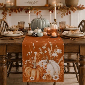 May include: A rectangular table runner in a burnt orange hue featuring a printed design of white and orange pumpkins surrounded by dried wildflowers and foliage. The runner is displayed on a wooden dining table set with plates and candles.