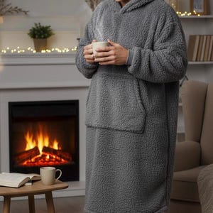 May include: A gray, oversized hooded blanket with a large front pocket. The cozy, plush blanket is worn by a person holding a mug. The background includes a fireplace, a side table, and a bookshelf, creating a warm, inviting atmosphere.