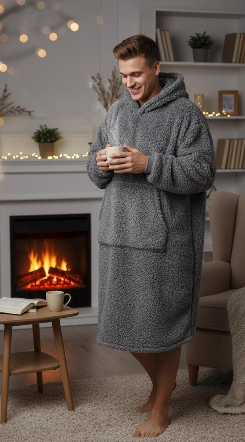 May include: A gray, oversized hooded blanket with a large front pocket. The cozy, plush blanket is worn by a person holding a mug. The background includes a fireplace, a side table, and a bookshelf, creating a warm, inviting atmosphere.