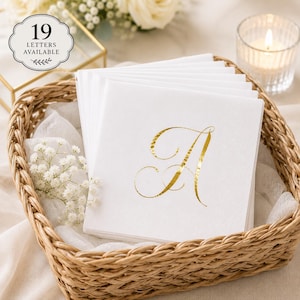 May include: A stack of square white paper napkins featuring a metallic gold foil letter A in a cursive script font. The napkins are displayed in a woven wicker basket. A circular badge in the corner displays the text 19 LETTERS AVAILABLE.