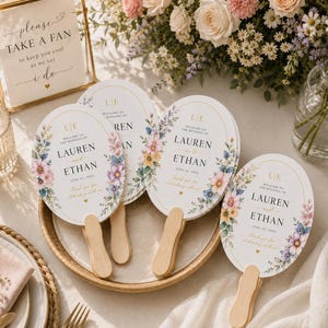 May include: Oval-shaped wedding fans with wooden handles, featuring floral watercolor illustrations in pink, blue, and yellow. The text reads: Welcome to the wedding of Lauren and Ethan, June 21, 2025, Thank you for celebrating with us, and the initials LE.
