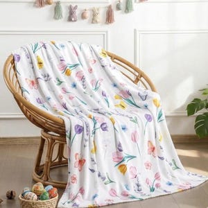 Puede incluir: Una manta floral blanca sobre una silla de mimbre. La manta presenta un estampado de coloridos tulipanes y mariposas. Una pequeña cesta de huevos de Pascua decorados está en el suelo. Una guirnalda decorativa con conejitos y borlas cuelga en el fondo.