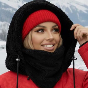 May include: A black hooded scarf and a red beanie. The scarf has a drawstring closure and a soft, textured appearance. The person in the image is wearing a red jacket. The background shows a snowy mountain scene.
