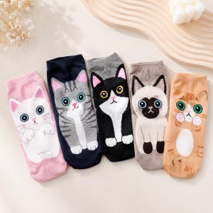 May include: Five pairs of ankle socks featuring cartoon cat designs. The socks are pink, navy blue, gray, light beige, and tan. Each sock displays a different feline face with large eyes, small ears, and paws positioned at the bottom of the design.