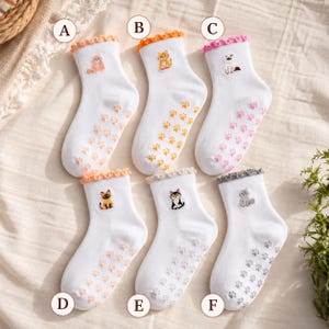 Puede incluir: Seis calcetines blancos de tobillo con pu&ntilde;os con volantes, cada uno con un peque&ntilde;o gato bordado y agarres de huellas de patas a juego en la suela. Los calcetines est&aacute;n etiquetados de la A a la F, con dise&ntilde;os de gatos en naranja, amarillo, rosa, marr&oacute;n, negro y gris.