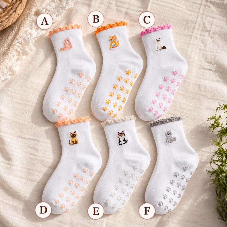 Puede incluir: Seis calcetines blancos de tobillo con pu&ntilde;os con volantes, cada uno con un peque&ntilde;o gato bordado y agarres de huellas de patas a juego en la suela. Los calcetines est&aacute;n etiquetados de la A a la F, con dise&ntilde;os de gatos en naranja, amarillo, rosa, marr&oacute;n, negro y gris.
