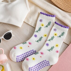 Könnte beinhalten: Ein Paar weiße, gerippte Socken mit einem kleinen Blumenmuster aus gelben, violetten und rosa Blüten sowie grünen Blättern. Die Socken haben einen violetten Rüschensaum am Bündchen und violette, rutschfeste Silikonnoppen an den Sohlen.