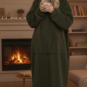 May include: A dark green hooded blanket with a large front pocket. The blanket is worn by a person holding a mug. The background includes a fireplace, a small table, and a bookshelf, creating a cozy, relaxed atmosphere.