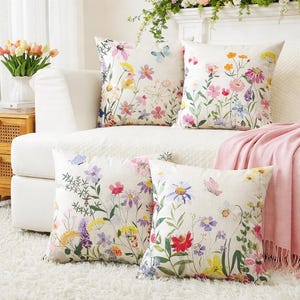 May include: Four square throw pillows with floral designs. The pillows feature a white background with colorful flowers and butterflies. The pillows are on a white couch with a pink blanket.