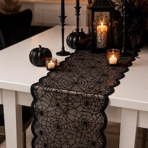 Peut inclure: Un chemin de table en dentelle noire orné d'un motif complexe de toile d'araignée avec des bords festonnés. Le chemin est posé sur une table blanche, accompagné de bougeoirs noirs, d'une lanterne noire, de petites citrouilles et de bougies.