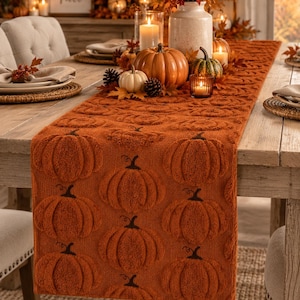 May include: A rectangular burnt orange table runner featuring a repeating raised texture pattern of pumpkins with dark brown stems. The fabric has a soft, tufted appearance and covers the length of a wooden dining table, creating a seasonal aesthetic.