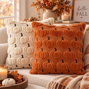 May include: Two square throw cushions featuring a repeating tufted pumpkin pattern. One cushion is cream-coloured with cream pumpkins, and the other is burnt orange with matching orange pumpkins. Both have a textured knit base and dark stems.