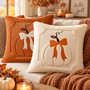 May include: Two square throw pillows with a textured corduroy border and a central plush pumpkin motif. One pillow is burnt orange with a white pumpkin and bow, while the other is cream with an orange pumpkin and bow, both featuring dark stems.