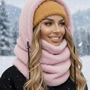 May include: A pink fleece hood and scarf combination with a mustard yellow knit beanie. The hood has drawstrings and a soft, textured appearance. The person is wearing a black sweatshirt with white text.