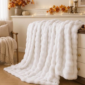 Puede incluir: Una manta blanca de felpa con un patrón texturizado de rayas verticales. La manta está colocada sobre el borde de un mueble de madera, mostrando su material suave, grueso y esponjoso con un color blanco brillante y uniforme en toda ella.