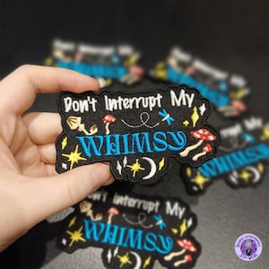 May include: A black embroidered patch with the text "Don't Interrupt My WHIMSY" in white and blue. The patch features whimsical designs like mushrooms, stars, and a crescent moon. The patch is held in a hand, with other patches in the background.