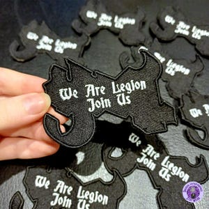 May include: Black embroidered patch with the white text "We Are Legion Join Us". The patch has a distinctive, stylized shape with curved edges. Several similar patches are visible in the background.
