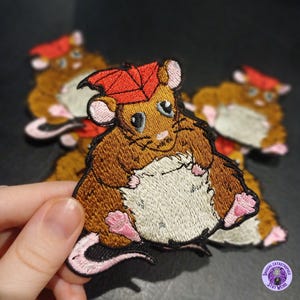 Könnte beinhalten: Gestickter Aufnäher mit einem Cartoon-Hamster, der einen roten Blatthut trägt. Der Hamster hat braunes Fell, einen weißen Bauch und rosa Pfoten und Ohren. Der Aufnäher ist mit detaillierten Stichen und einem skurrilen Stil gestaltet.