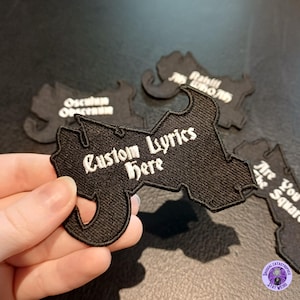 May include: Black embroidered patches with white text. One patch reads "Custom Lyrics Here." Other patches have text that is partially visible. The patches are in a unique shape. The patches are on a dark surface.