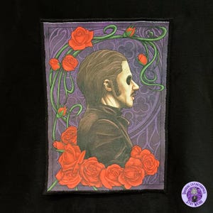 May include: A rectangular patch featuring a profile portrait of a figure with a painted face, surrounded by red roses and green vines against a purple background. The patch has a black border and a small circular logo that says "Unusual Catastrophes, Stay Weird."