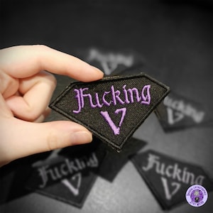 May include: A black diamond-shaped patch with purple embroidered text reading "Fucking 17". The patch is held up by a hand, with other patches visible in the background. A distinctive accessory.