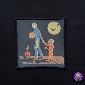 May include: A black and white embroidered patch featuring a dark-haired man in a blue shirt and jeans holding a jack-o'-lantern. A small, orange, stitched figure with a burlap sack head is holding a small jack-o'-lantern and walking alongside the man. The background is a dark blue with a full moon and a bat.