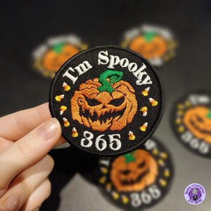 Puede incluir: Parche negro bordado con un diseño de calabaza de Halloween. El parche presenta el texto "I'm Spooky 365" en blanco, con detalles de caramelos de maíz. La calabaza es naranja con un tallo verde. Otros parches están en el fondo.
