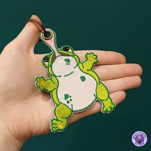 May include: Embroidered frog patch with a green and white design, featuring a long pink tongue. The patch has a key ring attachment. The frog is in a playful pose, with its arms and legs outstretched.