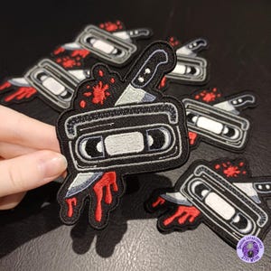 May include: Embroidered patch depicting a black VHS tape with a silver knife and red blood splatter. The patch has a black border and is designed for application on clothing or accessories.