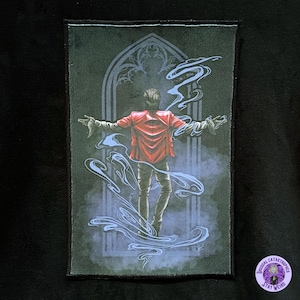 May include: A rectangular patch featuring an illustration of a person in a red jacket with arms outstretched, standing before a gothic archway. The person is surrounded by blue, swirling smoke. The patch has a black border and a small, circular logo that says "Unusual Catastrophes Stay Weird."