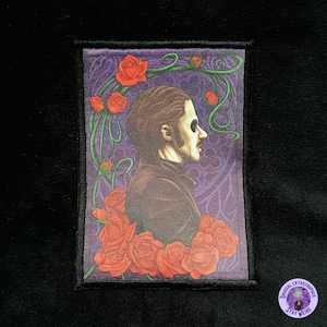 Könnte beinhalten: Rechteckiger Aufnäher mit einem Profilporträt einer Person mit bemaltem Gesicht, umgeben von roten Rosen und grünen Ranken auf lila Hintergrund. Der Aufnäher hat einen schwarzen Rand und ein kleines rundes Logo mit "Unusual Catastrophes, Stay Weird."