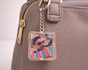 May include: Square keychain with a photo of two people kissing, attached to a silver chain and keyring. The keychain is hanging from a gray purse. The photo shows a man wearing sunglasses and a woman in a striped shirt.
