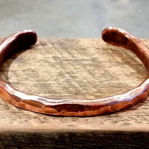 May include: A hammered copper bracelet with a smooth, rounded finish. The bracelet is made from a single piece of copper and has a simple, minimalist design.