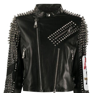 May include: Black leather jacket with silver metal spikes on the shoulders and sleeves. The jacket features a zip closure, multiple pockets, and a small patch with a design on the right sleeve. The jacket has a round collar.